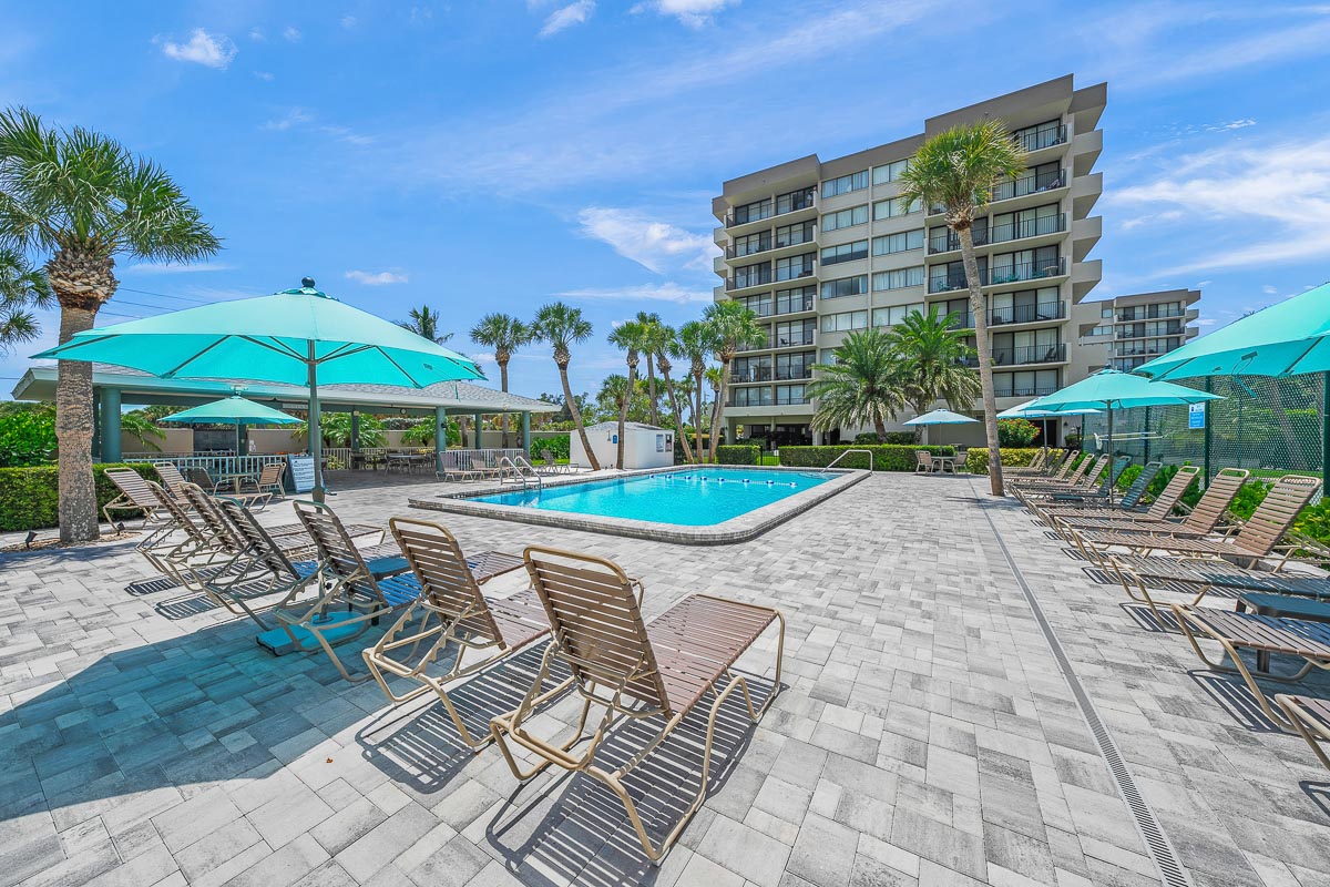 A pool area with lounge chairs, umbrellas, and palm trees in front of a multi-story building.